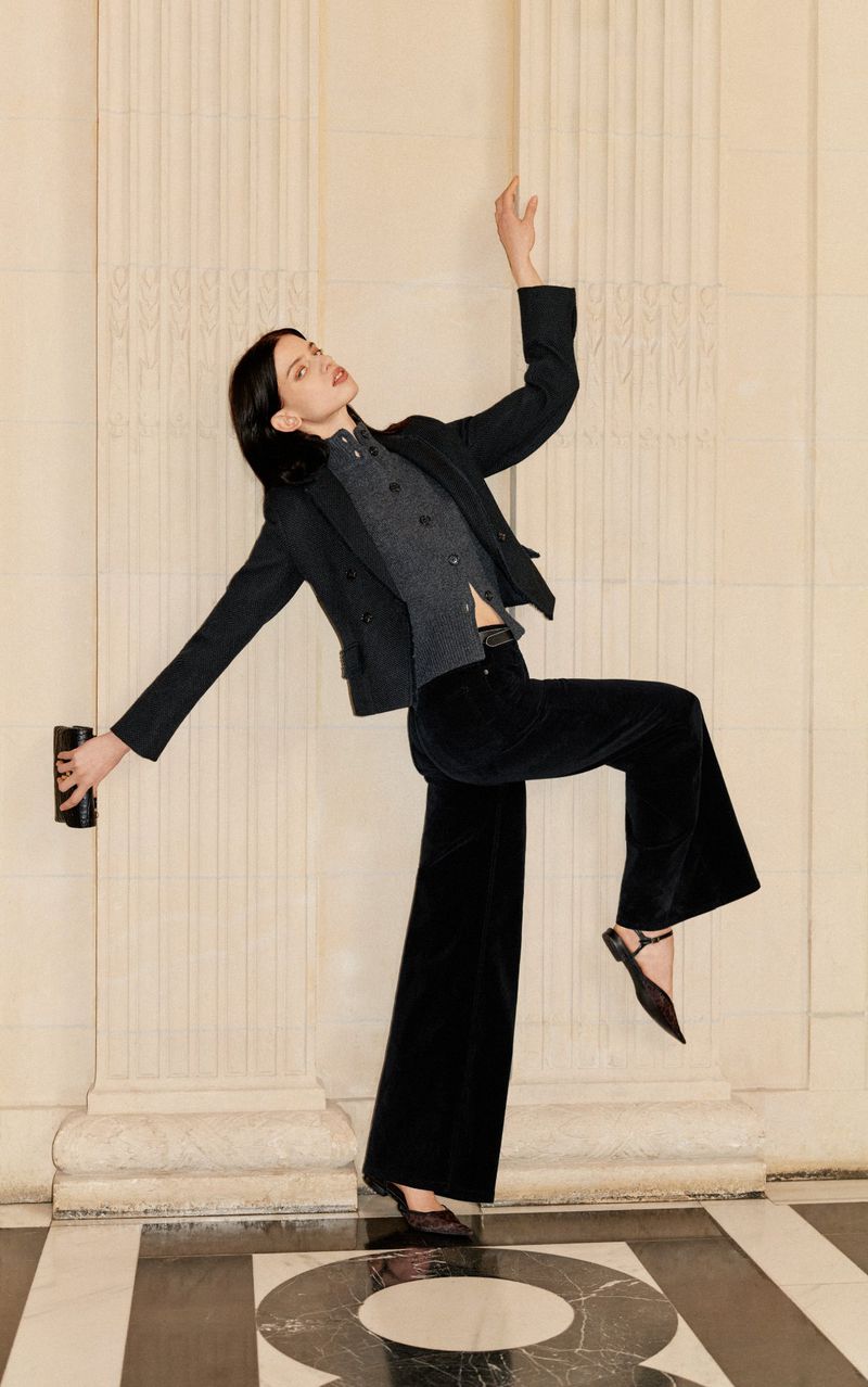 Adult female model poses in a black blazer, vest, and wide-leg trousers with decorative columns and a patterned floor in the background.