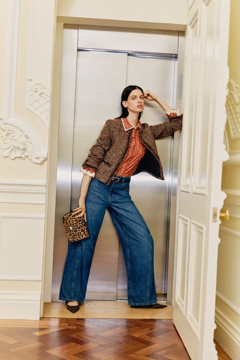 A stylish adult woman in a patterned top and wide-leg jeans poses in an elevator doorway with a leopard print purse, showcasing fashion.