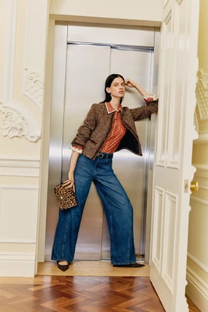 A stylish adult woman in a patterned top and wide-leg jeans poses in an elevator doorway with a leopard print purse, showcasing fashion.