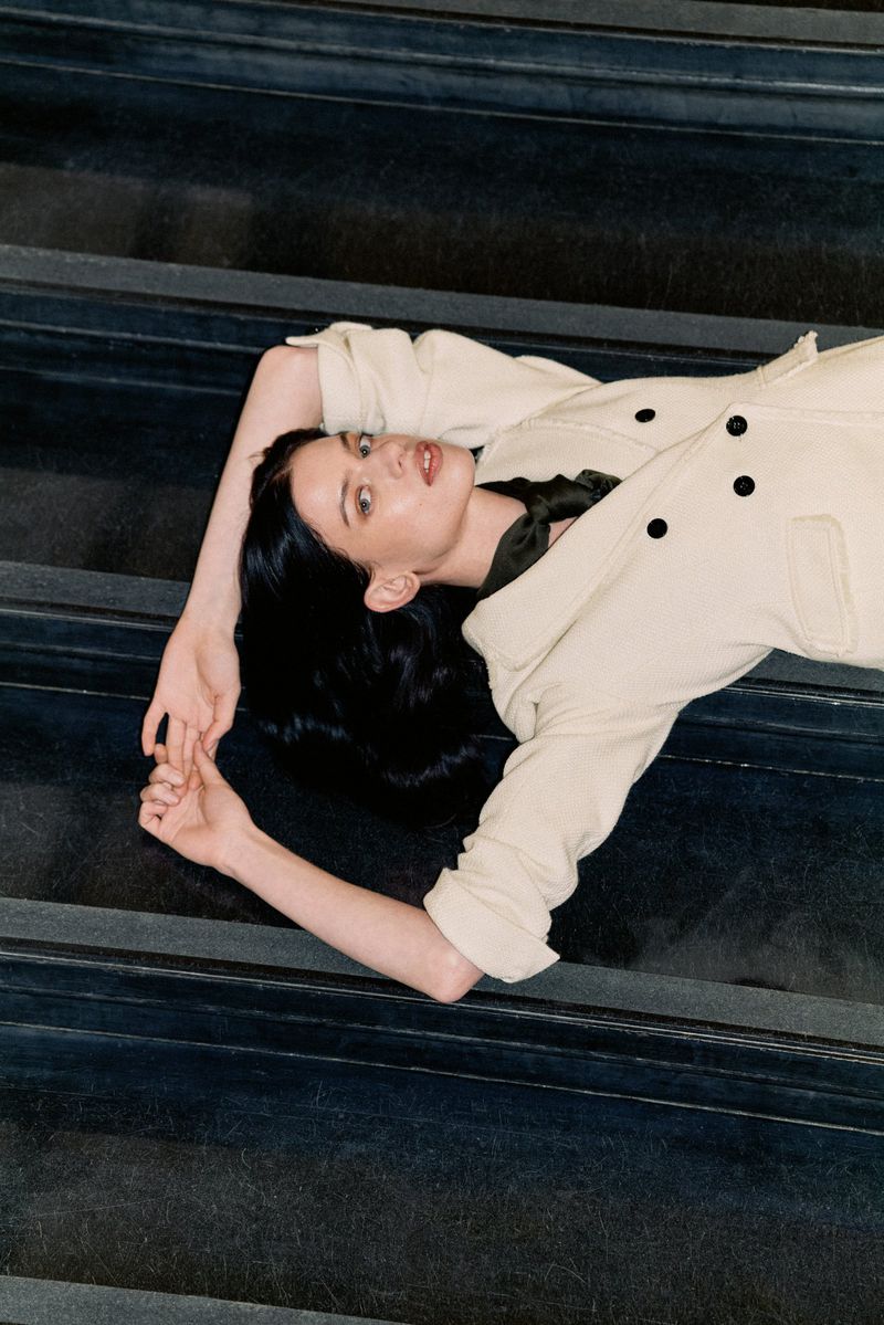 A woman with long black hair and a white coat poses laying on dark stairs, looking upwards in a fashion or editorial style image.