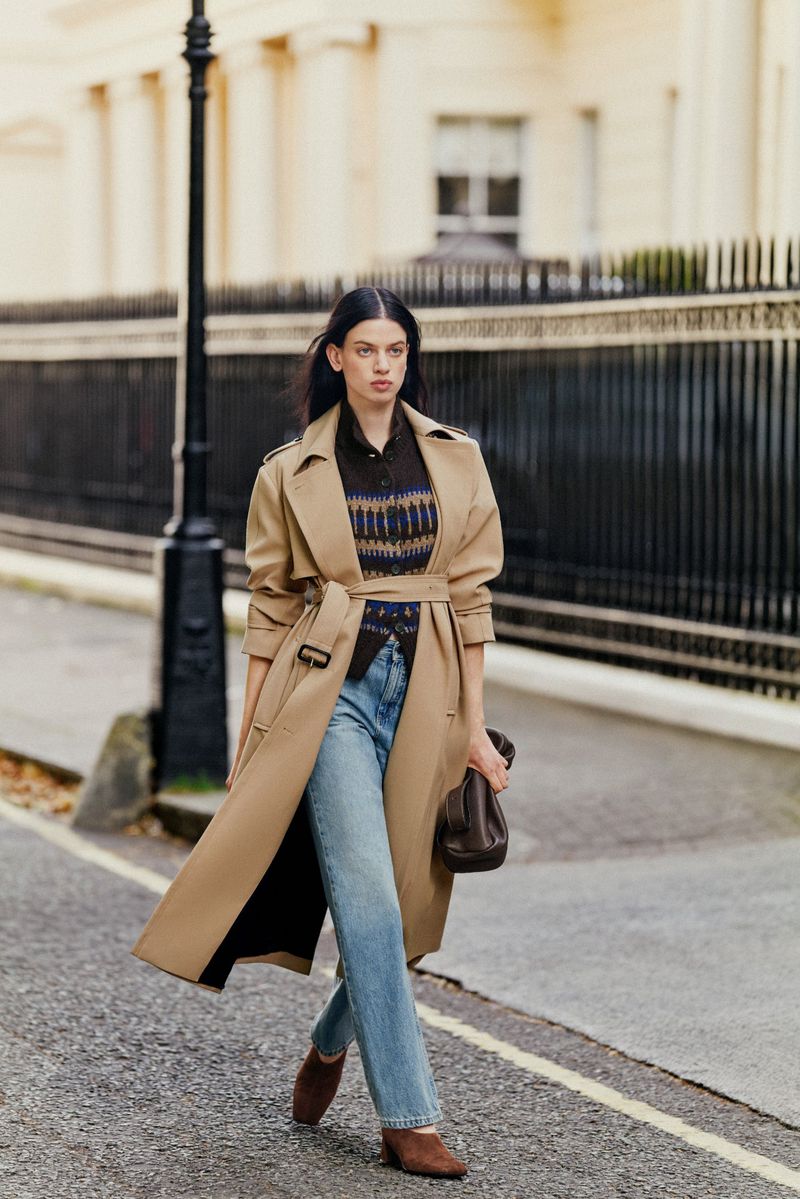 Model Lily McMenamy wearing a camel trench coat and blue jeans walking past a classic town house exterior.