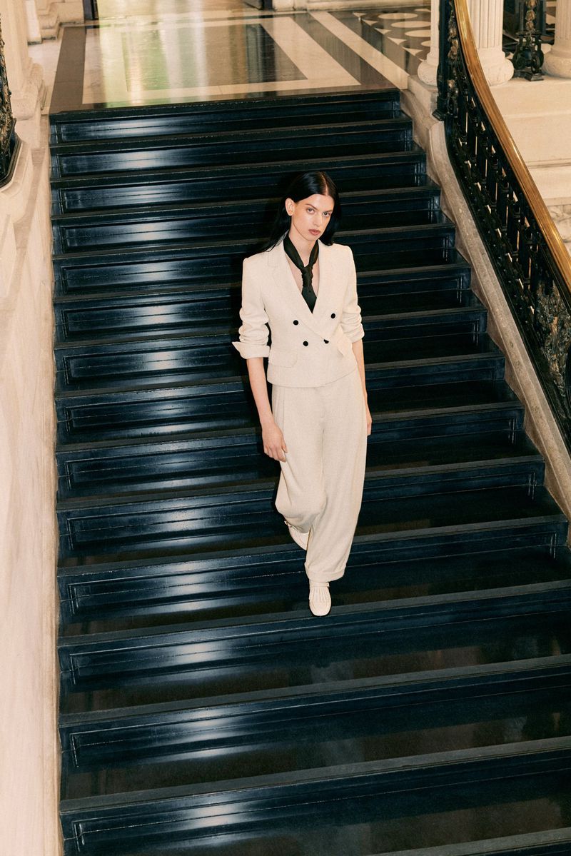Adult woman in an off-white suit descends a black staircase. She wears a black scarf and white shoes, photographed indoors.