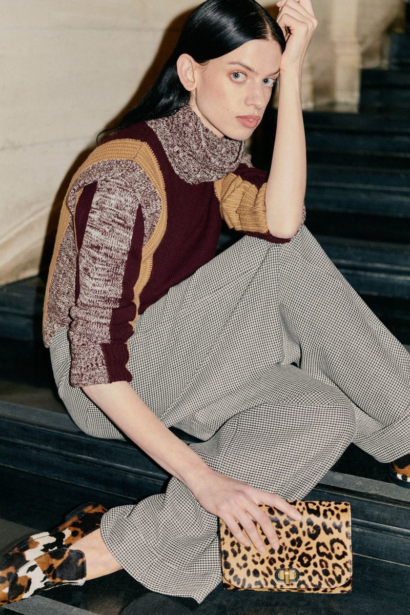 Model Lily McMenamy seated on dark steps wearing a color-blocked turtleneck sweater and houndstooth trousers