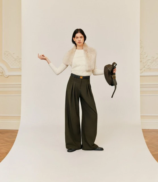 Fashionable adult woman in studio wearing cream sweater, olive green trousers and black shoes and holding dark green bag with white backdrop.
