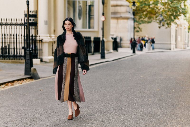 A stylish woman in a fur-trimmed jacket and striped skirt confidently walks down a European street with blurred pedestrians behind her.