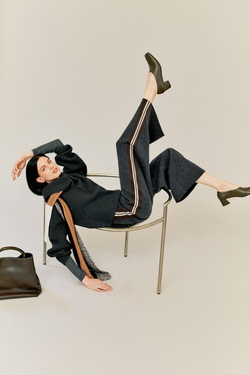 Woman in gray sweater and striped pants reclines on a chair in a fashion studio with a black bag. Elegant pose and neutral backdrop.