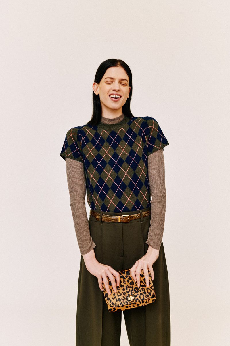A woman with dark hair smiles and poses wearing a green and navy argyle sweater, olive-green pants, and holding a leopard-print purse.