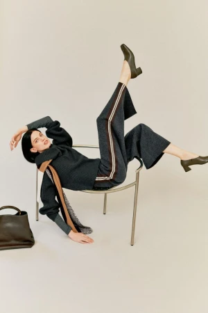Model in grey knit sweater and trousers with side stripes posing playfully on a metal chair against a plain light background
