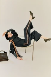 Model in grey knit sweater and trousers with side stripes posing playfully on a metal chair against a plain light background