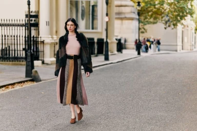 Model walking on a city street wearing a color-blocked skirt, pink knit top, and fur-trimmed jacket.