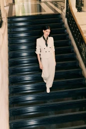 Model Lily McMenamy descending dark wooden staircase wearing cream tailored trouser suit and dark necktie for ME+EM