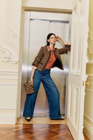 Model leaning against a metal elevator door wearing a tweed jacket, printed blouse, wide-leg jeans, and holding a leopard print handbag.
