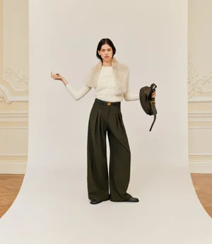 Model Lily McMenamy posing in an off-white sweater, fur scarf, and dark trousers against a white seamless background