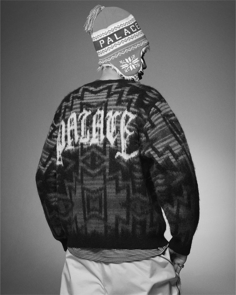 A person wearing a Palace brand sweater and winter hat is seen from the back against a gray background in a studio shot.