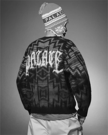 A person wearing a Palace brand sweater and winter hat is seen from the back against a gray background in a studio shot.