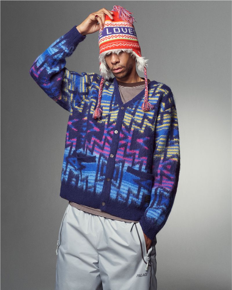 Man in patterned cardigan and beanie poses indoors against a gray backdrop, creating a fashion-forward editorial shot.