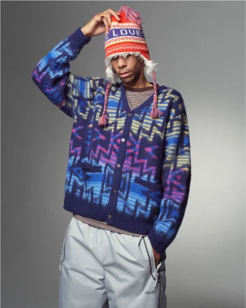 Man in patterned cardigan and beanie poses indoors against a gray backdrop, creating a fashion-forward editorial shot.