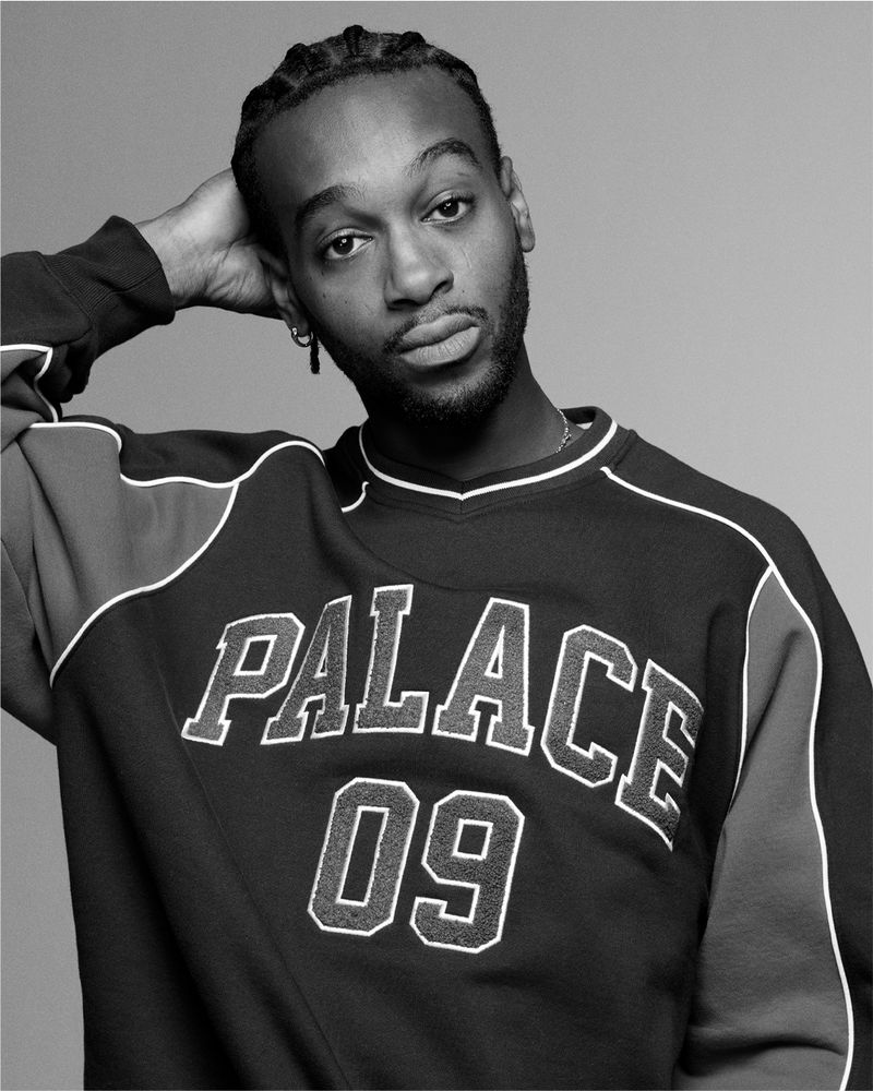 Portrait of a young African American man in a 'Palace 09' sweatshirt, hand on head, against a grey background, photographed indoors.