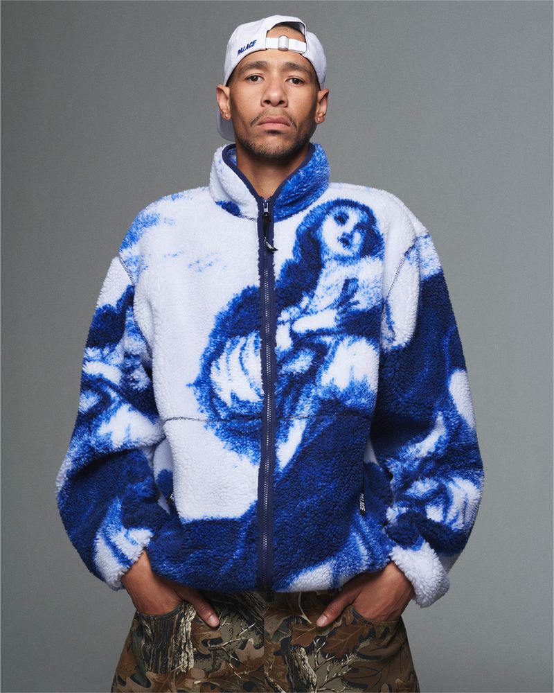 Adult male poses in a studio wearing a baseball cap, graphic sherpa jacket with camouflage pants against a solid grey background.