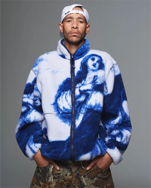 Adult male poses in a studio wearing a baseball cap, graphic sherpa jacket with camouflage pants against a solid grey background.
