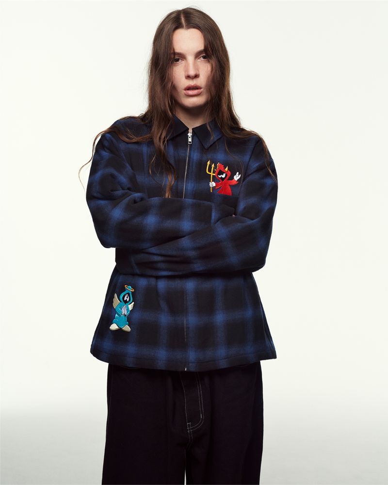 Adult woman in a medium studio shot, wearing a blue plaid jacket with embroidered devil and angel characters, and black pants.