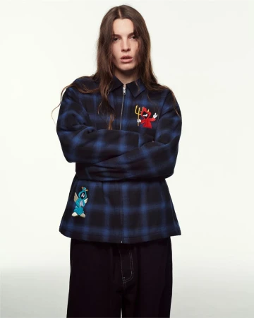 Adult woman in a medium studio shot, wearing a blue plaid jacket with embroidered devil and angel characters, and black pants.