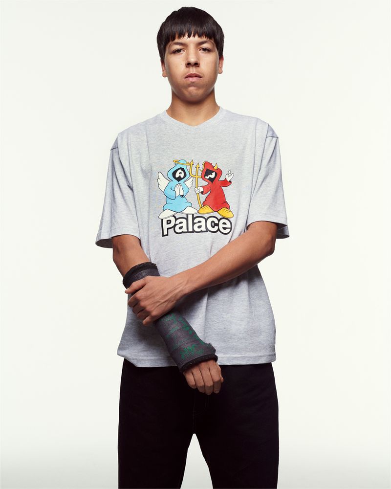 Young adult poses in gray Palace T-shirt with cartoon angel and devil graphic, black pants, and a forearm cast, against a white backdrop.