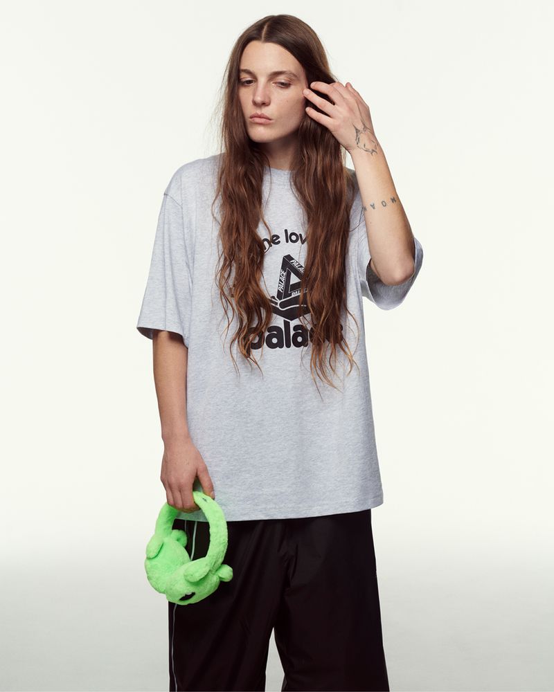A person with long hair is in a studio holding a green plush frog purse with a gray t-shirt and black shorts in a studio shot.