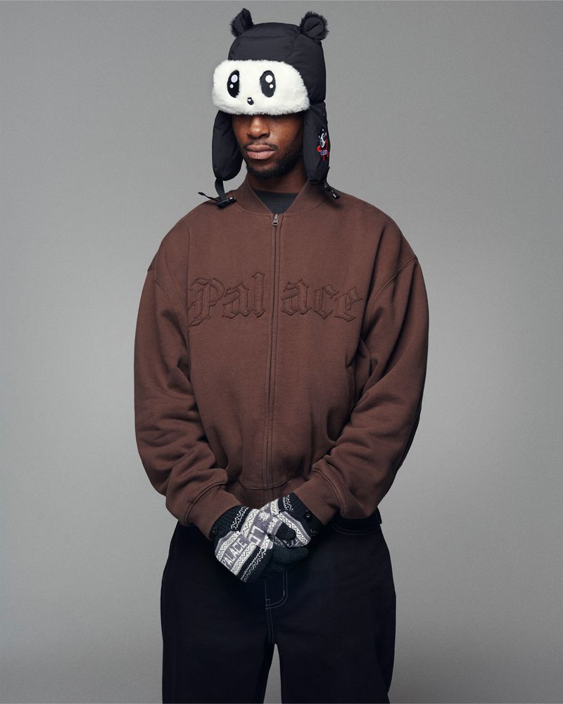 An African-American male posing indoors, wearing a brown bomber jacket, black pants, novelty hat, and gloves against a neutral background.