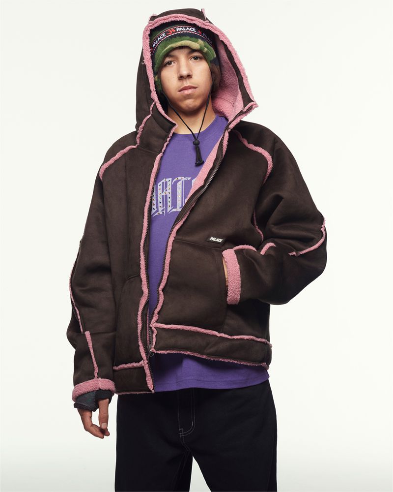 A young person wearing a brown hooded jacket with pink lining, purple t-shirt, and patterned beanie stands in a medium studio shot.