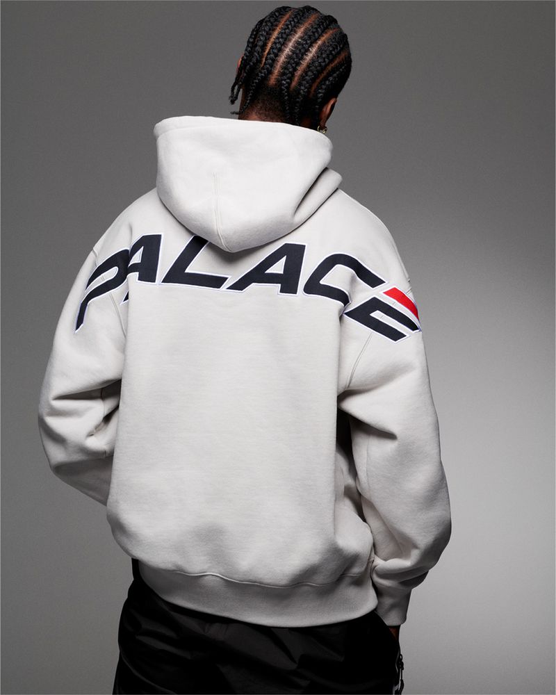 Back view of an adult male wearing a white Palace hoodie with black accents and braided hair, set against a gray background.