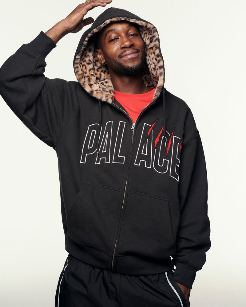 A smiling Black man wearing a black zip-up hoodie with a leopard-print lined hood poses in front of a neutral-colored background.