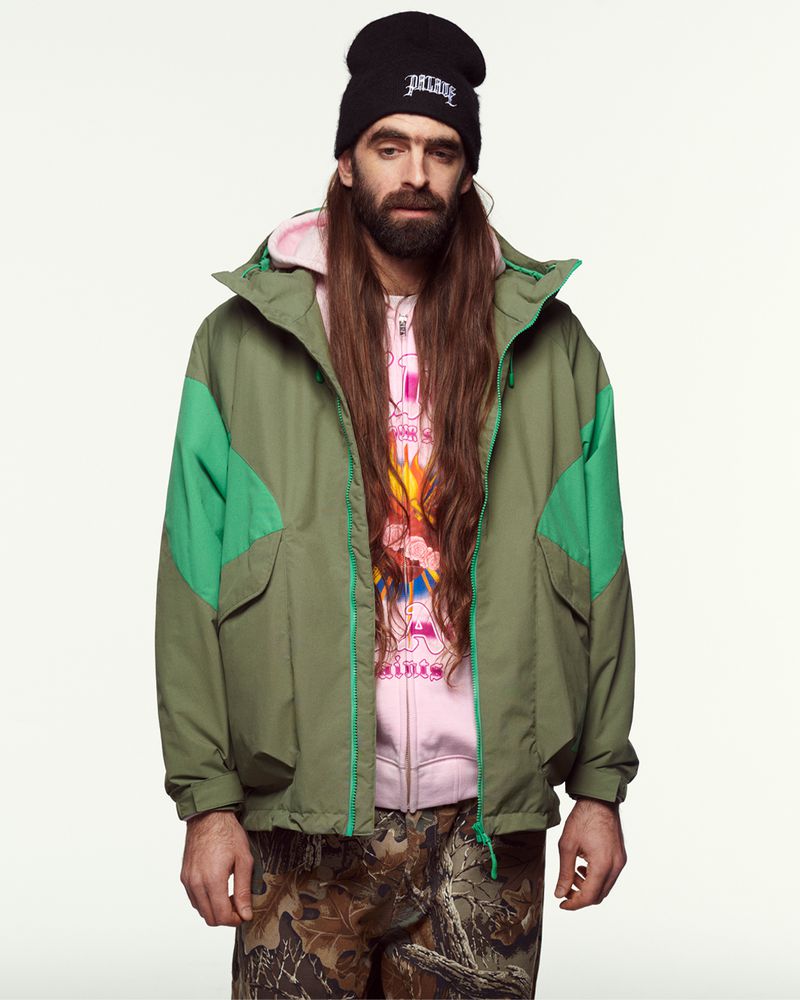 Adult male with long hair wearing a black beanie, green jacket, pink hooded sweatshirt, and camouflage pants against a white background.