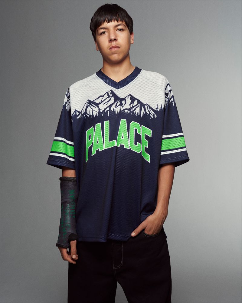 Young man in navy 'Palace' jersey with mountain graphic posing in a studio with arm cast, black pants, and looking at camera.