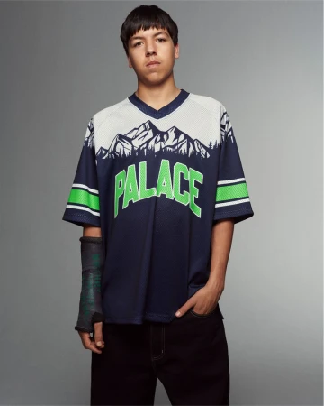 Young man in navy 'Palace' jersey with mountain graphic posing in a studio with arm cast, black pants, and looking at camera.