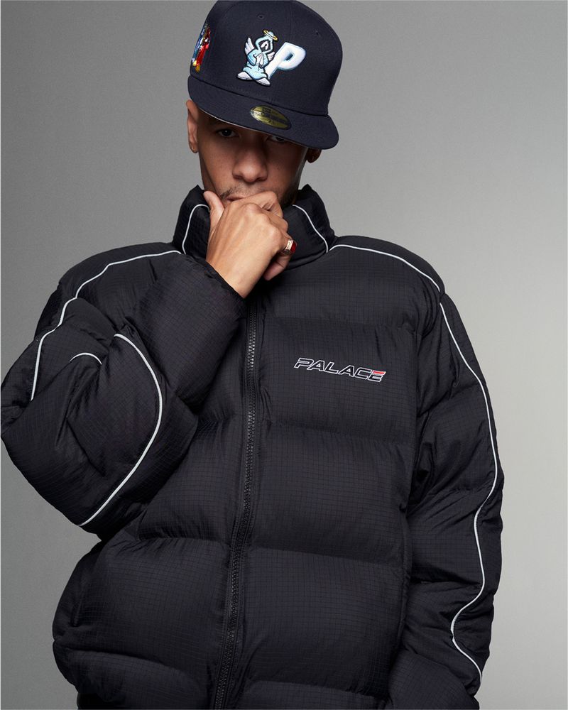 Young adult poses in a black puffer jacket with white piping and a Palace baseball cap against a neutral grey studio backdrop.