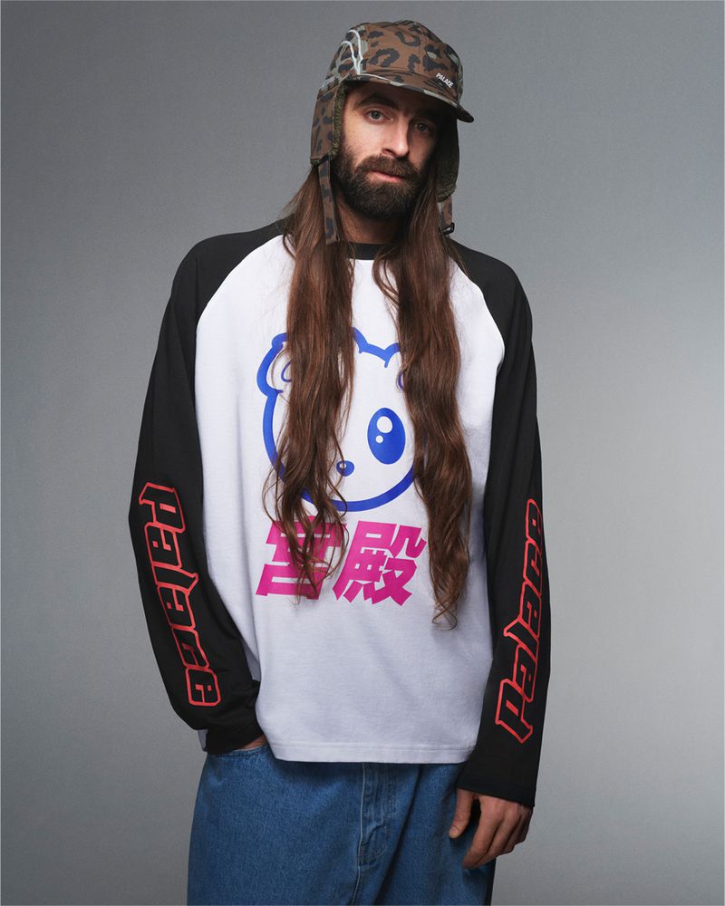 Adult male with long brown hair wears a novelty hat and graphic long-sleeve shirt with Japanese text in a studio setting.