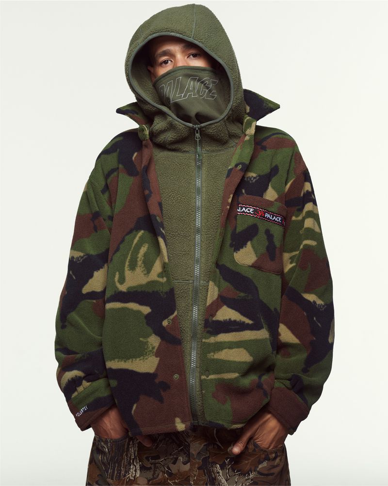 Adult male in a fleece zip-up jacket with a camo pattern, wearing a balaclava, and a hood, standing indoors in a studio setting.