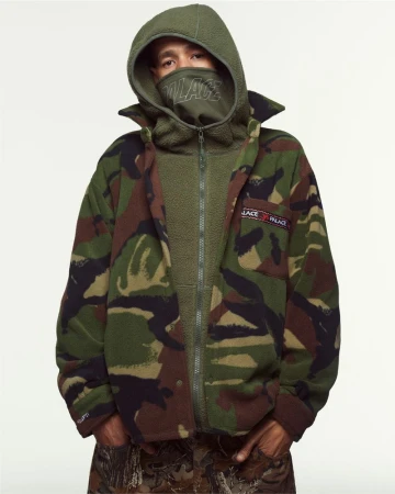 Adult male in a fleece zip-up jacket with a camo pattern, wearing a balaclava, and a hood, standing indoors in a studio setting.