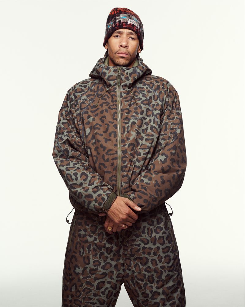 Man in patterned jumpsuit and beanie stands against a white backdrop in a studio portrait, creating a striking fashion statement.