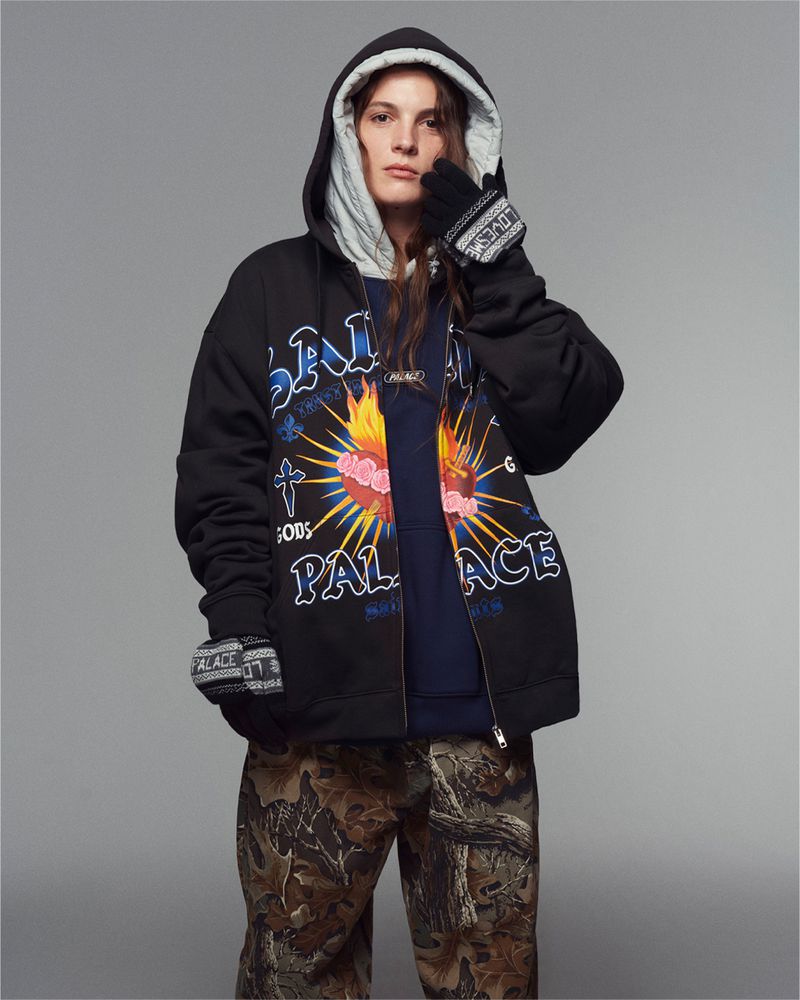 Person in a black hooded sweatshirt with graphic designs, paired with camouflage pants, against a gray backdrop in a studio setting.