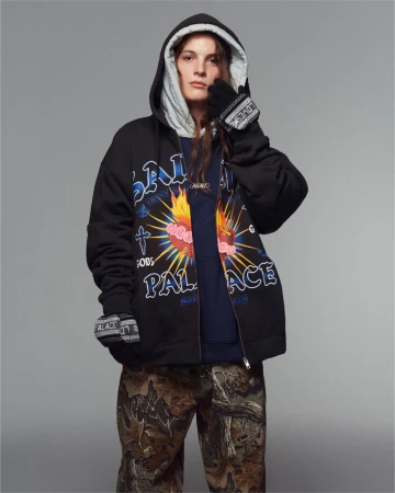 Person in a black hooded sweatshirt with graphic designs, paired with camouflage pants, against a gray backdrop in a studio setting.