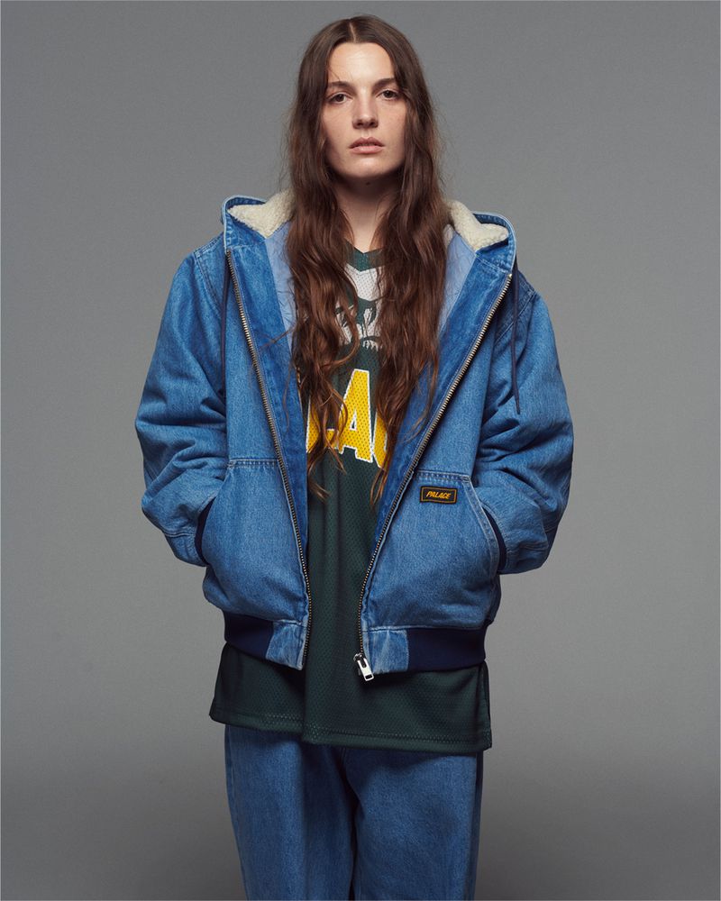 Studio shot of a woman with long brown hair wearing a Palace branded denim jacket and jeans against a neutral grey backdrop.