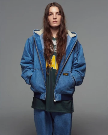 Studio shot of a woman with long brown hair wearing a Palace branded denim jacket and jeans against a neutral grey backdrop.