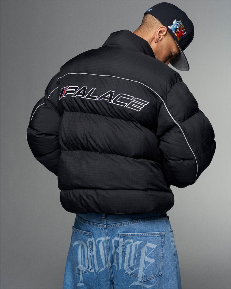 Person wearing a black Palace puffer jacket and light blue Palace logo jeans, accessorized with a baseball cap, against a grey background.
