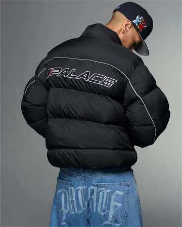 Person wearing a black Palace puffer jacket and light blue Palace logo jeans, accessorized with a baseball cap, against a grey background.