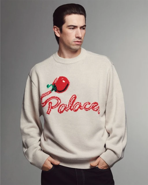 Studio shot of a man wearing a knit sweater with a cherry and the word 'Palace' across the front, hands in pockets, against a gray background.