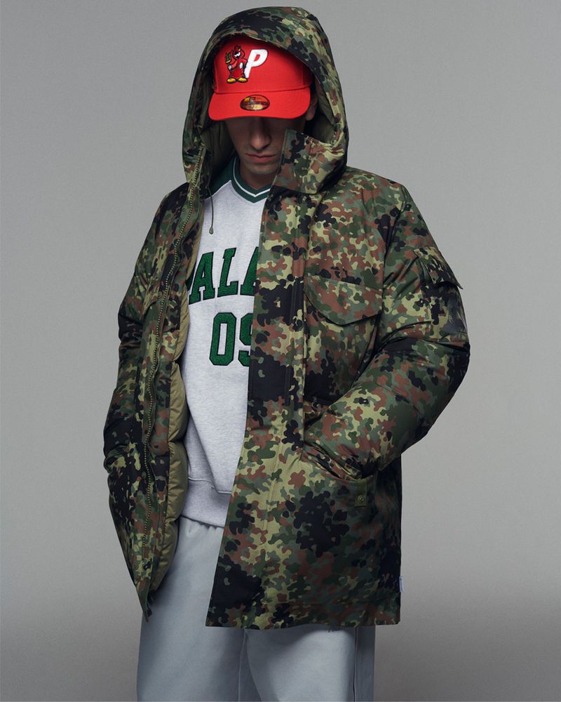 Man in camo hooded puffer jacket, sweatshirt, sweatpants, and red ball cap posing in a photo studio with a neutral grey background.