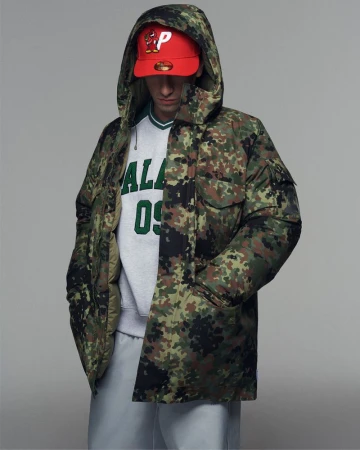 Man in camo hooded puffer jacket, sweatshirt, sweatpants, and red ball cap posing in a photo studio with a neutral grey background.