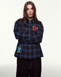 Model with long brown hair wearing a blue plaid jacket with embroidered patches against a white background for PALACE Holiday 25.